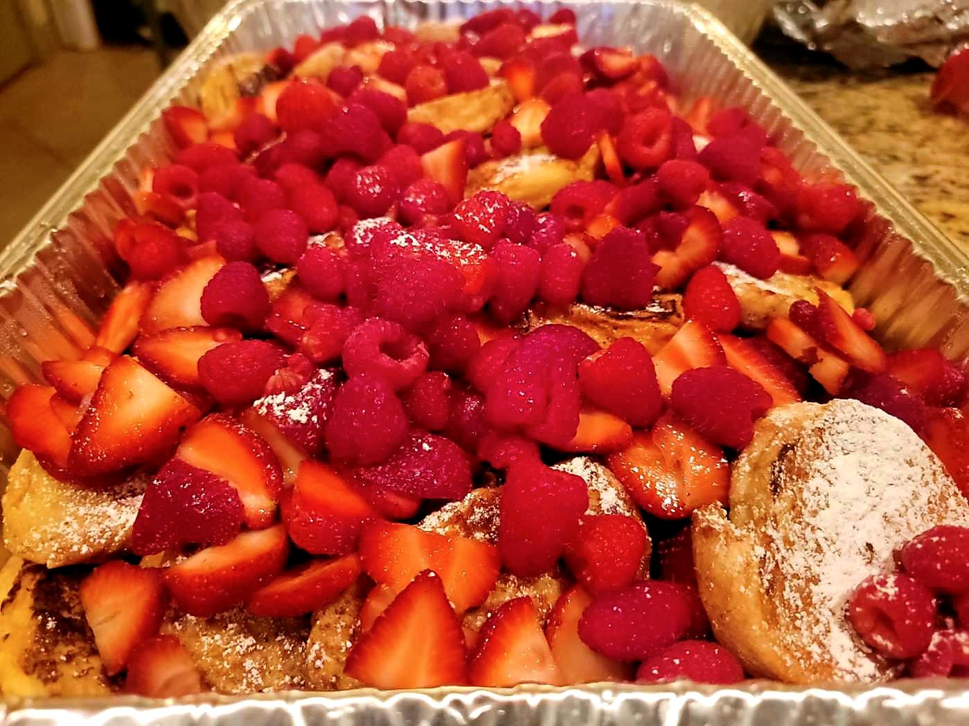 A catering tray full of golden baked treats topped with strawberries, raspberries, and powdered sugar.
