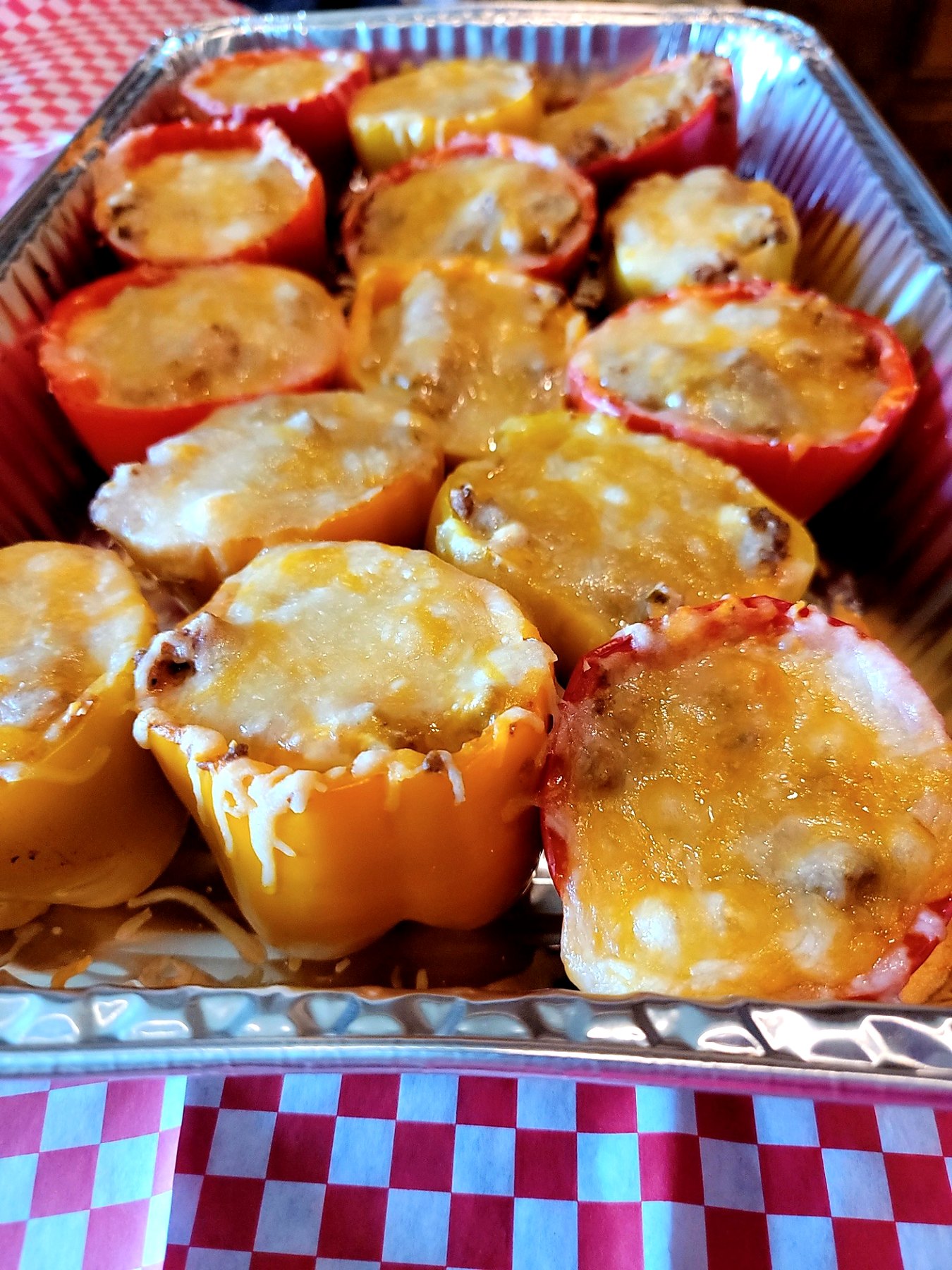 Red and yellow stuffed peppers with a melted cheese topping in a foil catering tray.